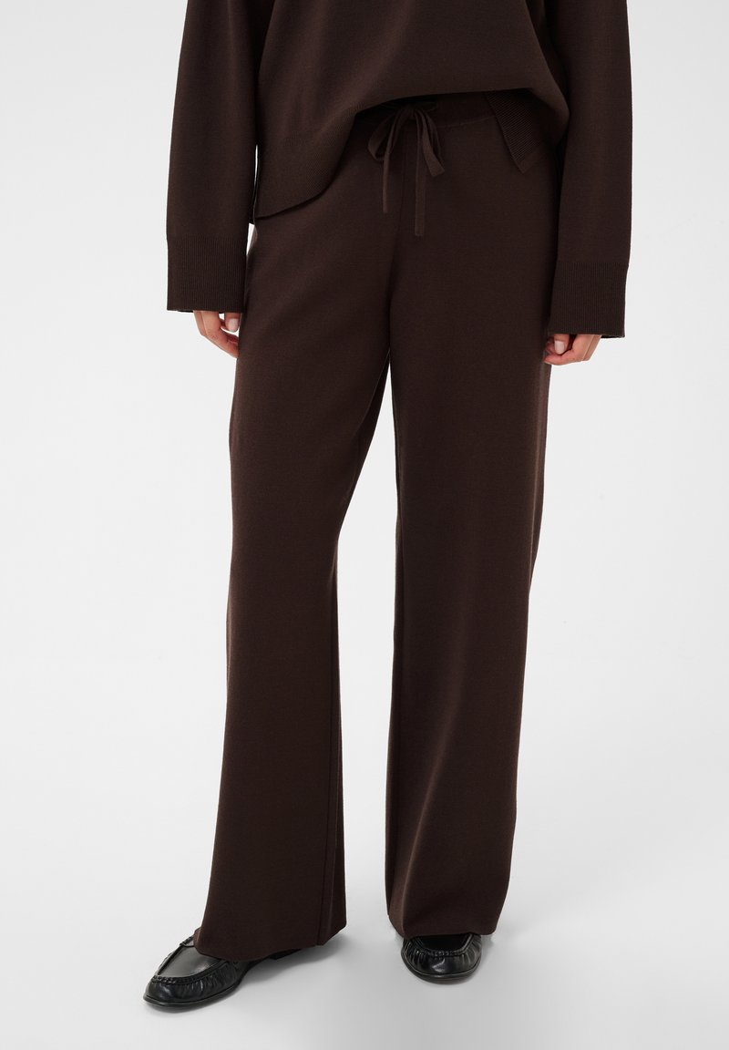 Personne portant un pantalon large marron foncé avec taille à cordon et un haut à manches longues assorti, associé à des chaussures plates noires.