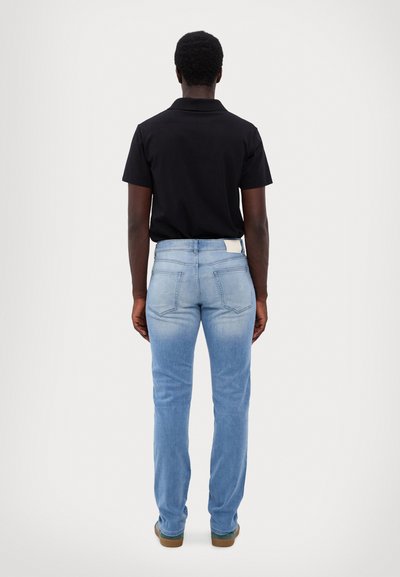 Jean en denim bleu, coupe droite, délavé clair, poches arrière, et une étiquette blanche à la taille, porté avec un polo noir.