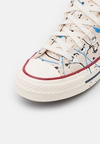 Plátěné tenisky s béžovým svrškem, na kterém jsou černé a modré skvrny. Bílý gumový špička a vínový akcent kolem podešve.