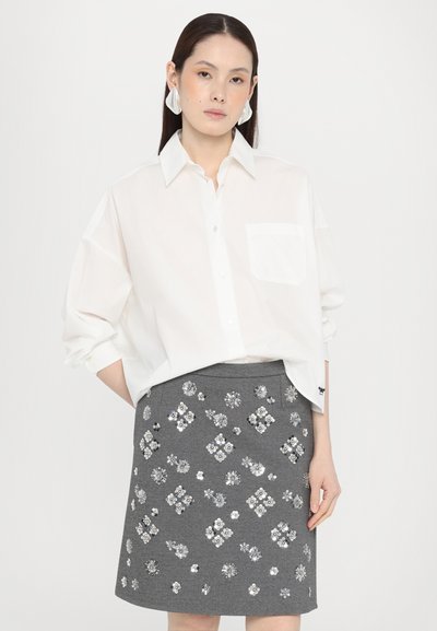 Hvit skjorte med knapper, med en enkelt brystlomme, kombinert med et grått skjørt som har utsmykkede blomsterdesign i sølvtoner.