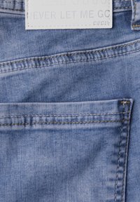 Ljust blå denimtyg med en synlig ficka och kontrasterande sömmar. En vit etikett visar "DENIM NEVER LET ME GO."