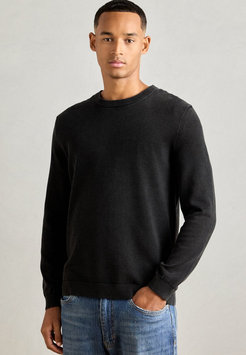 Schwarzer Strickpullover mit Rundhalsausschnitt und Rippbündchen, kombiniert mit hellblauen Jeans. Strukturierter Stoff, figurbetonte Form, lässiges Design.
