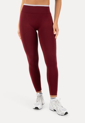 Persona que lleva leggings granate de cintura alta combinados con zapatos deportivos blancos y calcetines blancos de tobillo con marca, de pie frente a un fondo liso.