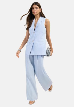 Nainen vaaleansinisessä hihattomassa bleiserissä ja leveälahkeisissa housuissa kävelee, pitäen hopeista kytkintä, pukeutuneena hopeiseen rannekoruun ja valkoisiin korkokenkiin.