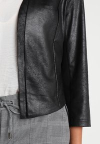 Veste en cuir noir texturée portée par-dessus un haut blanc avec un pantalon à carreaux noir et blanc noué à la taille.