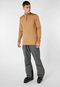 Brun lynlåset langærmet top med sidelægningsdetaljer, parret med grå skibukser og sorte skistøvler, båret af en stående mandlig model.