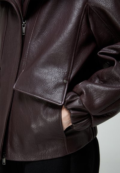 Tmavohnedá kožená bunda so zipsom vpredu, detailom s klopou na vrecku a elastickým lemom na rukáve, kde je čiastočne zasunutá ruka vo vrecku.