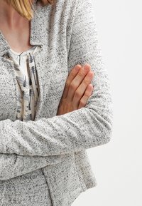Chaqueta gris con estampado y tejido texturizado, con cuello tipo chal y mangas largas. Debajo lleva una blusa de color claro con un patrón floral.