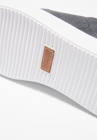 Svart texturerad sneakersula med en vit gummibotten. Har en tan läderetikett inbäddad på sidan, med räfflade mönster för grepp.