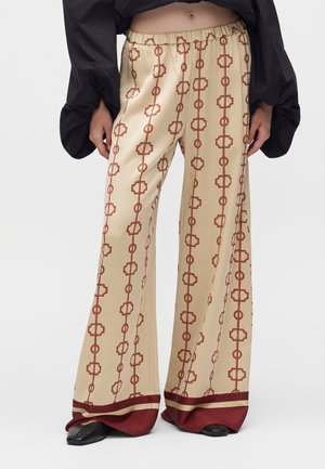 Beige bukser med vide ben med røde geometriske lodrette mønstre og mørkerød kant, båret med sort top og sorte spidse sko.