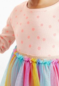 Rosa ribbad klänning med orange prickar, med en regnbågsfärgad tylskjol med volanglager och ett guldigt elastiskt midjeband.