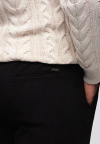Gros plan d'une personne portant un pull en maille torsadée crème et un pantalon noir avec une poche arrière et une main dans la poche avant.
