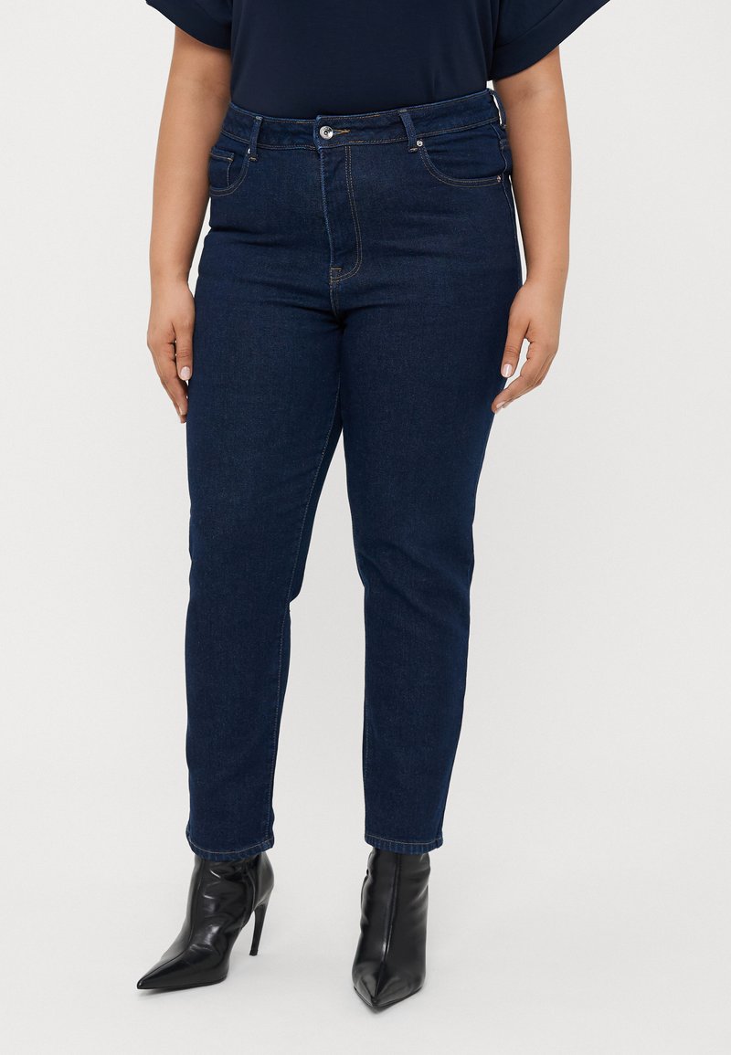 Tamno plave traperice ravnog kroja, s patentnim zatvaračem, pet džepova i vidljivim šavovima. Uparene s crnim špicastim čizmama do gležnja.