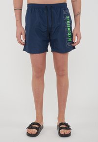 Marine zwemshorts met een tailleband met trekkoord, voorzien van verticale tekst in groen en wit aan de zijkant. Gladde, lichte stof.