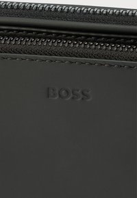 Portafoglio in pelle nera con texture liscia, dotato di chiusura a zip e logo "BOSS" in rilievo sul davanti. Design semplice e minimalista.