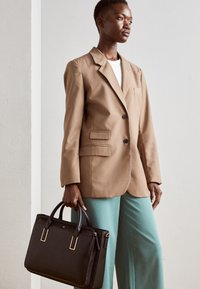 Brun blazer med hakkede revers, sort håndtaske med gulddetaljer, kombineret med teal bukser med vide ben. Glat stof og struktureret design.