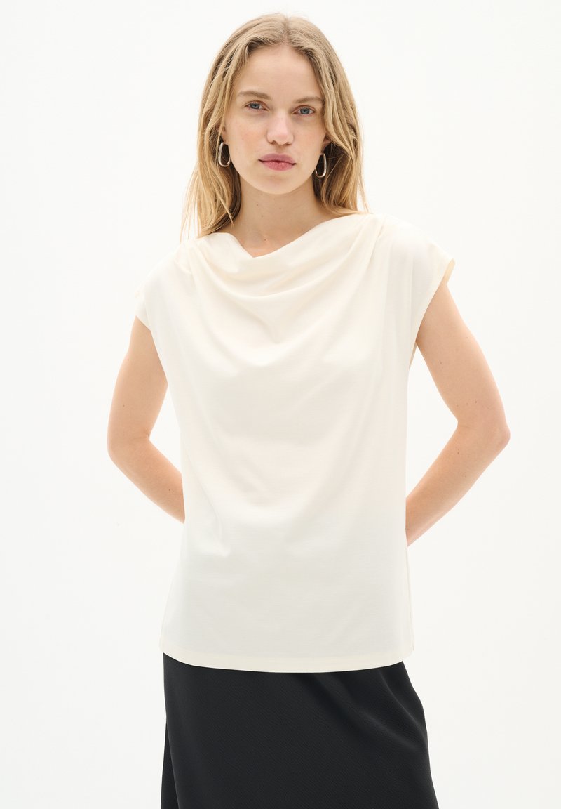 Haut drapé crème avec des manches courtes, fabriqué en tissu doux. Il présente un col rond et une coupe décontractée, associé à une jupe noire.