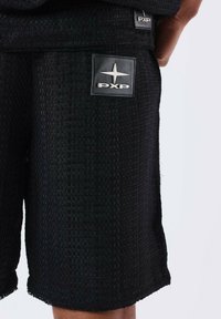 Pantaloni corti neri in tessuto intrecciato con una finitura testurizzata, un orlo grezzo e un'etichetta quadrata nera con un design di stella argentata.