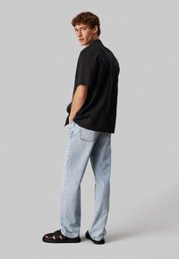 Joven vestido con una camisa negra de manga corta, jeans azul claro y sandalias negras, de pie con las manos en los pockets, mirando por encima del hombro.