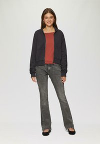 Zwarte ritsjack over een roestkleurige top, gecombineerd met grijze flared jeans en zwarte flats. Glad materiaal met ribbels aan de manchetten en tailleband.