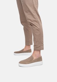 Zapatos de ante marrón tipo slip-on con suela de goma blanca, usados con pantalones cortos beige. Los zapatos presentan un sencillo bordado y una textura suave.