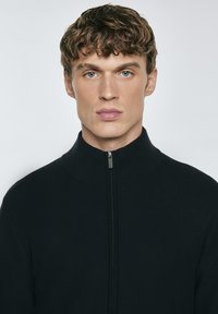 Svart zip-up stickad tröja med hög krage, som har en texturerad yta och en smal passform. Enkel och ren design, utan synliga mönster eller detaljer.