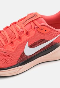 Nike trkačka cipela s koralnim mrežastim gornjim dijelom, teksturiranim uzorkom, bijelim swooshom, vezicama i crnom čizmom s ružičastim detaljima.