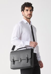 Malette en cuir noir avec un design structuré, fermetures à double boucle et une bandoulière amovible. Texture lisse avec des accents épurés.