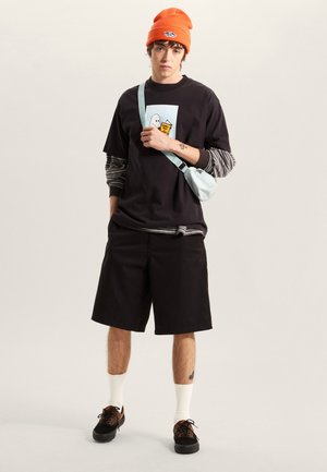 Schwarze Baumwollshorts, übergroßes schwarzes T-Shirt mit Grafik, darunter ein langärmliges, gestreiftes Hemd, orangefarbene Mütze und hellblaue Umhängetasche.