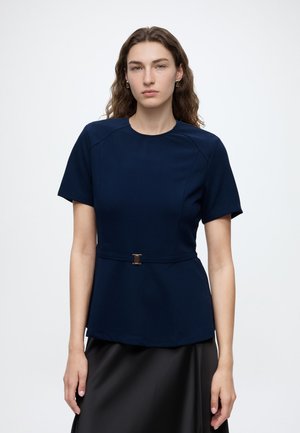 Femme aux cheveux bruns ondulés porte un haut bleu marine à manches courtes avec une ceinture fine et une jupe noire sur un fond clair uni.