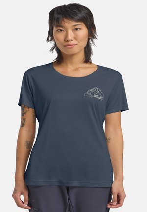 Junge Frau mit kurzen dunklen Haaren, die ein dunkelblaues Jack Wolfskin T-Shirt und dunkle Hosen trägt, steht entspannt mit den Armen.