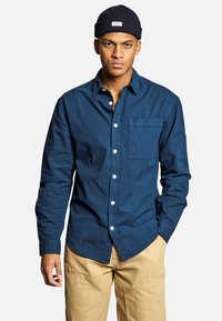 Camicia button-up blu navy con taschino sul petto e polsini ripiegati, realizzata in tessuto morbido, abbinata a pantaloni beige chiaro.