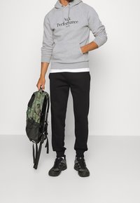 Homme portant un sweat à capuche gris "Peak Performance", un pantalon de survêtement noir, des baskets noires, et tenant un sac à dos camouflage vert par sa poignée.