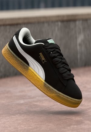 Basket Puma Suede noire avec bande de logo blanche et semelle jaune texturée, capturée en plein air sur un fond extérieur.