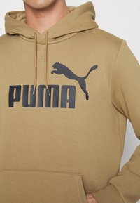 Felpa color khaki con tasca frontale a marsupio, caratterizzata da un grande logo nero "PUMA" e una grafica di un puma nero in salto. Materiale in morbido misto cotone.