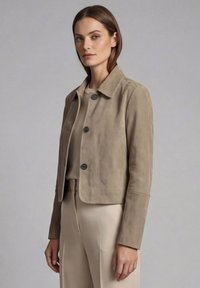 Kvinde iført en taupe croppet ruskindjakke over en beige top og beige højttaljede bukser mod en ensfarvet grå baggrund.