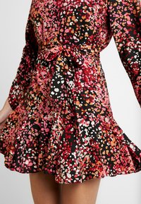 Femme portant une robe évasée à manches longues avec un motif floral abstrait coloré nouée à la taille avec une ceinture assortie.