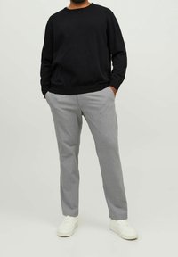 Homme portant une chemise noire à manches longues, un pantalon gris clair à fines rayures, des baskets blanches, debout les mains dans les poches sur fond blanc.