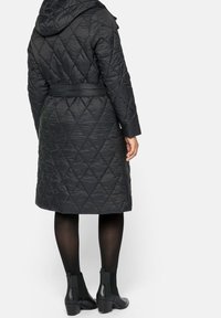 Abrigo largo acolchado negro con capucha, cintura ceñida y superficie texturizada; combinado con botas quitadas negras y medias opacas.