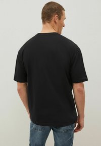 T-shirt in cotone nero con maniche corte, vestibilità rilassata e scollo rotondo, visto dalla parte posteriore. Indossato con jeans in denim blu.