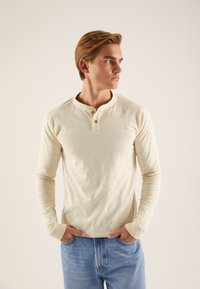Majica Henley z dolgimi rokavi v bež barvi, z okroglim ovratnikom, zapenjanjem na tri gumbe in rebrastimi manšetami. Mehka tkanina z gladko teksturo.
