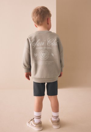 Een jong kind staat met de rug naar ons toe, draagt een grijze trui met de tekst "Safari Club", donkere korte broek, witte sokken en beige sneakers op een neutrale achtergrond.