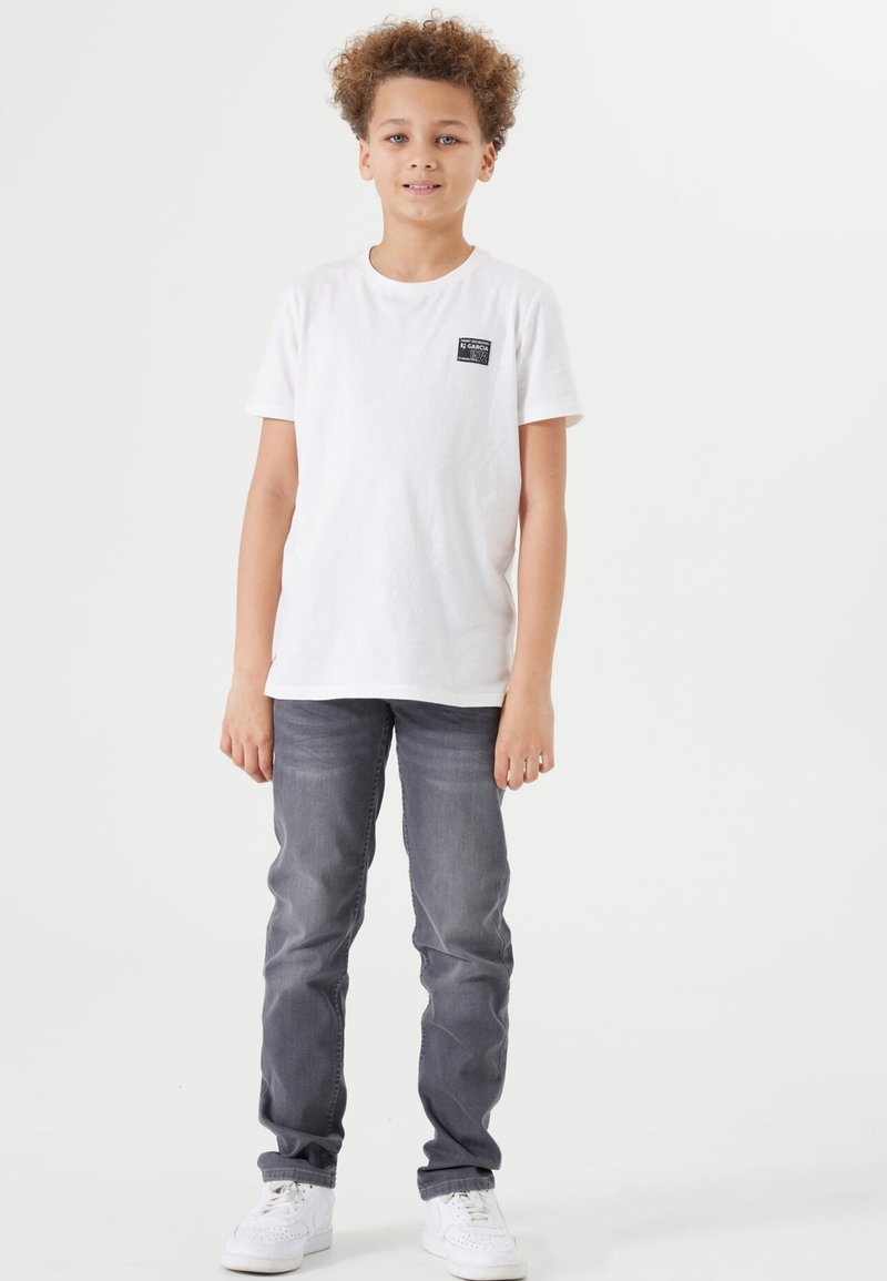 Witte katoenen T-shirt met klein zwart logo, gecombineerd met grijze jeans en witte sneakers; model staat tegen een neutrale achtergrond.
