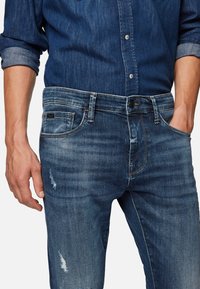 Faded blue denim jeans with a slim fit, distressed detailing, contrasting stitching, and a buttoned waistband.