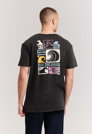 Homme portant un t-shirt noir avec des motifs ondulés colorés et le texte « Pro Test » et « Ride the Waves » au dos, debout sur un fond uni.
