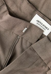 Close-up of brown pants with zipper and metal hook, showing a white label with "JACK & JONES PANTS STUDIO" text.