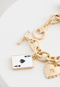 Zlatý náramek s řetízkem, na kterém je přívěsek s hracími kartami s černým srdcem a dvěma černými píky, vedle přívěsku ve tvaru srdce s diamantovým akcentem.