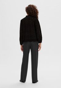 Blusa nera in pizzo con maniche lunghe, orlo ondulato e motivi intricati, abbinata a pantaloni grigio scuro, vista da dietro.