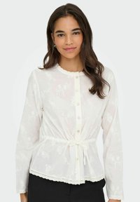 Blouse blanche à manches longues, décolleté festonné, taille nouée et motif floral brodé. Comprend une fermeture à boutons et une finition en dentelle.