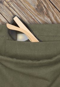 Sötét lencséjű napszemüveg fa karokkal, részben egy olajzöld szövet zsebében látható. Texturált szövet, varrás részletekkel.